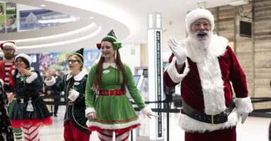 Man in white beard and Santa suit smiles and wavws with women in red and green elf outfits at mall entrance