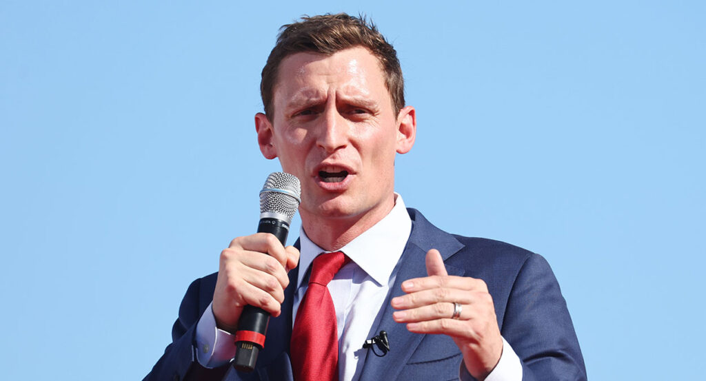 Blake Masters in a blue suit with a red tie speaks with a microphone in his right hand