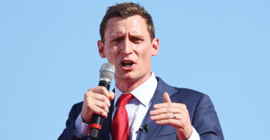 Blake Masters in a blue suit with a red tie speaks with a microphone in his right hand