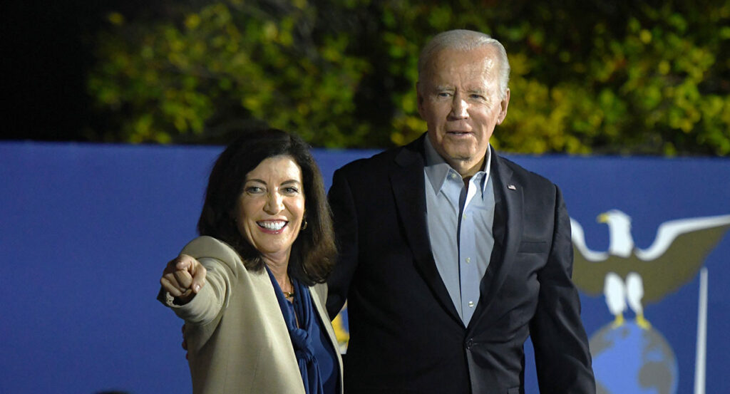 Black-haired woman in tan suit points as white-haired man in black suit stares blankly.