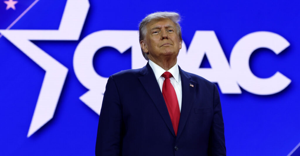 Trump in a suit in front of the CPAC logo