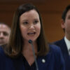 Ashley Moody in a blue suit speaks in front of Ron DeSantis in a blue suit.