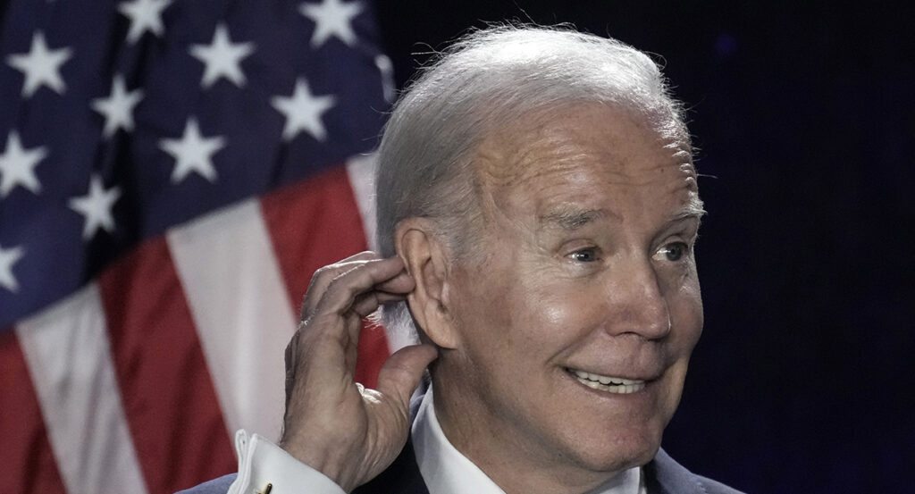 Joe Biden scratching ear in front of an American flag