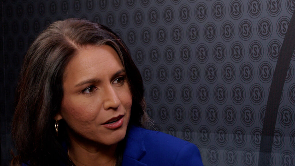 Tulsi Gabbard in a blue coat speaks in front of The Daily Signal logo