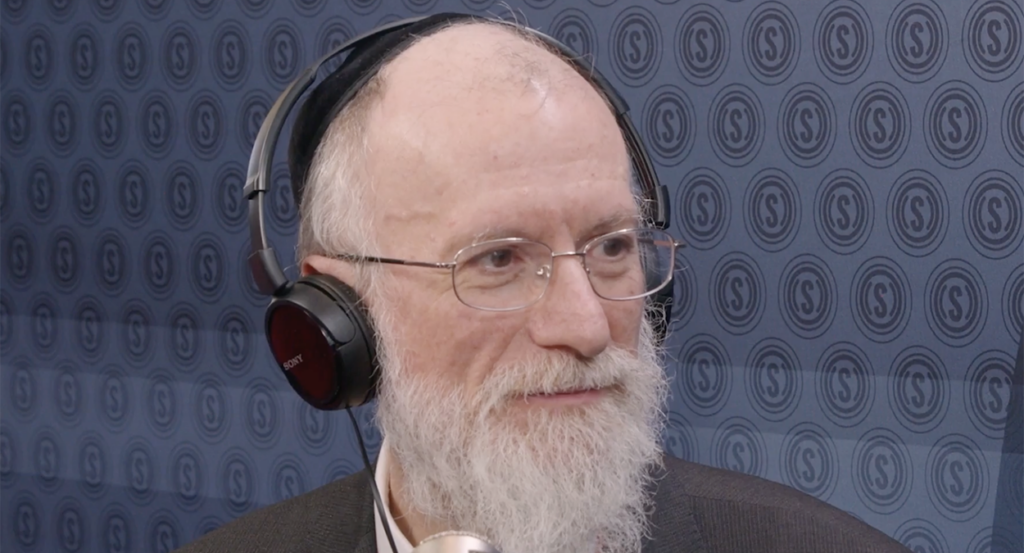 Rabbi Yaakov Menken with a white beard, glasses, and headphones in front of The Daily Signal logo