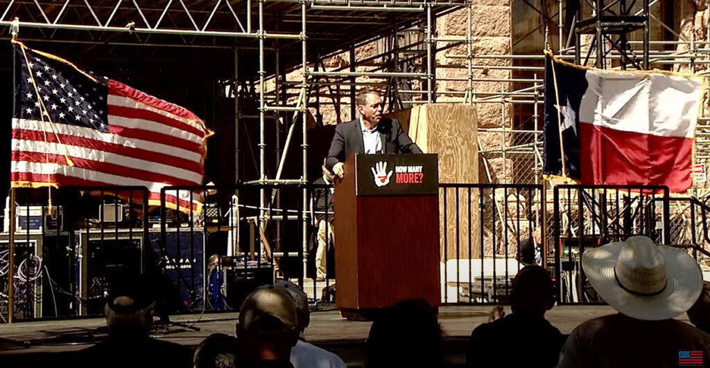 Mark Meckler speaks on a stage outside the Texas Capitol building with an American flag and a Texas flag behind him.