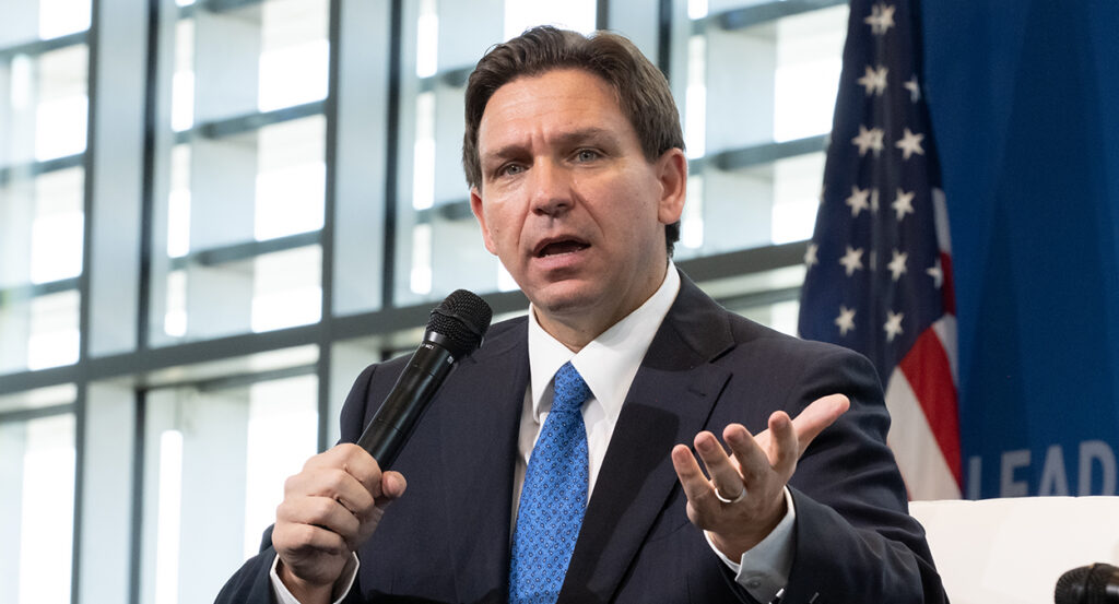 Florida Gov. Ron DeSantis in a black suit with a blue tie in front of an American flag