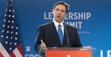 Ron DeSantis in a black suit with a blue tie in front of an American Flag and a blue Leadership Summit background with a Heritage Foundation logo