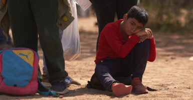 A young boy wearing a red shirt sits on the ground as he waits to be processed by Border Patrol.