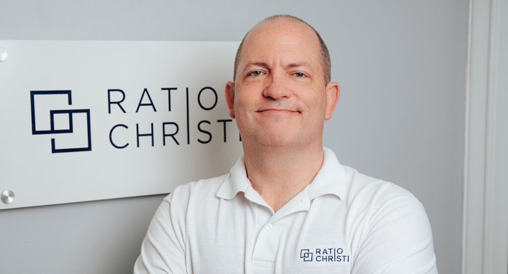 Corey Miller, wearing a white polo shirt reading "Ratio Christi" stands in front of the Ratio Christi office