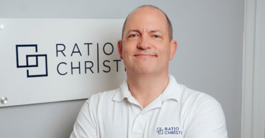 Corey Miller, wearing a white polo shirt reading "Ratio Christi" stands in front of the Ratio Christi office
