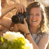Emilee Carpenter smiles while pointing a camera at a bride holding flowers