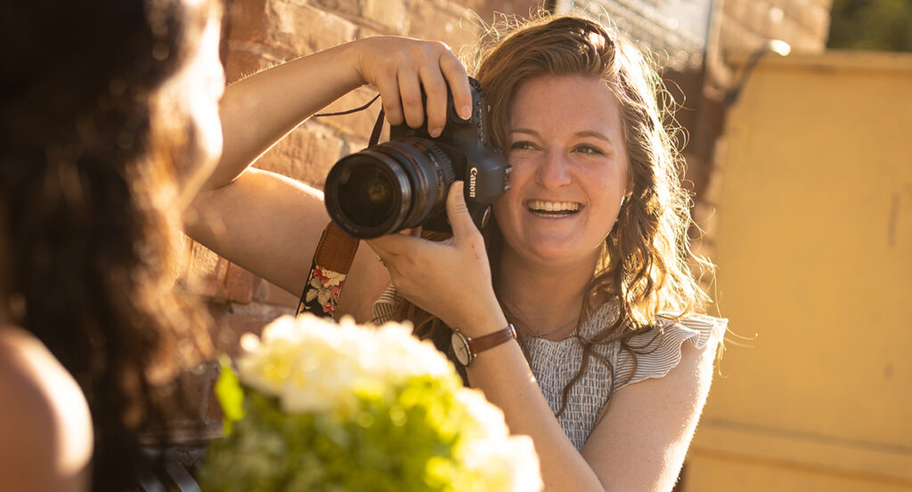 Emilee Carpenter smiles while pointing a camera at a bride holding flowers
