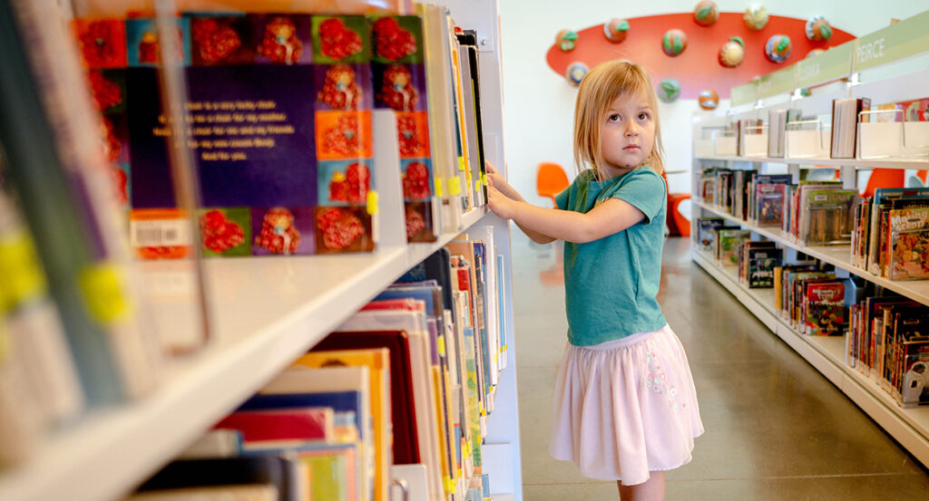 Elementary school girl in a grey shirt and white skirt peruses books in a library