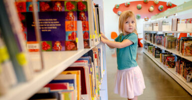 Elementary school girl in a grey shirt and white skirt peruses books in a library