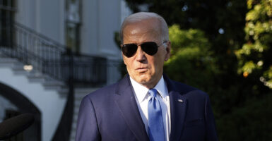Joe Biden stands with dark sunglasses in a blue suit in front of the White House
