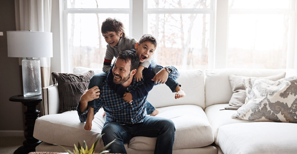 A father plays with his two sons on a white living room couch.