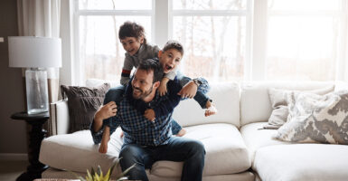A father plays with his two sons on a white living room couch.