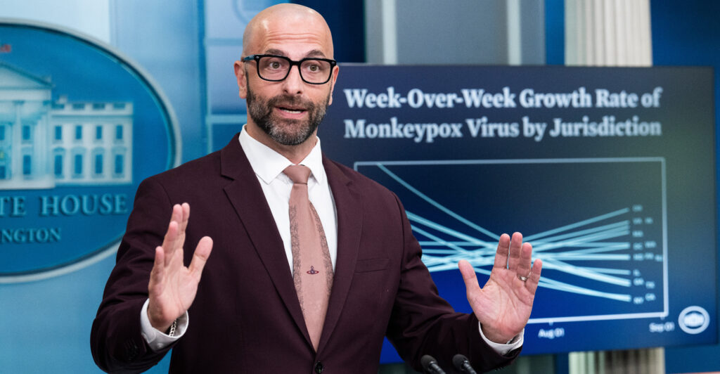 Demetre Daskalakis holds both of his hands up while speaking at a White House briefing. He stands in front of a blue White House emblem and TV screen showing statistics about Monkeypox.