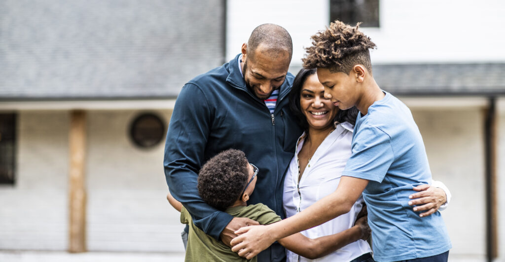 Portrait of a black father, mother, and two sons hugging in front of residential home.