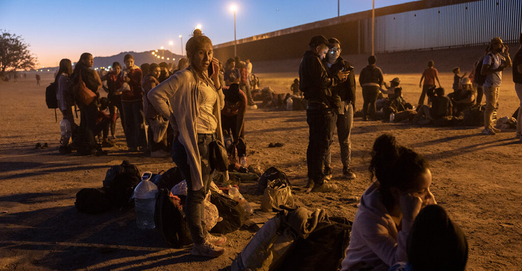 A group of illegal aliens sit and stand close to the border wall at dusk.