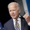 Joe Biden in a suit with an American flag pin holds a black mask in his left hand.