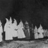 Men in white KKK hoods stand behind a burning cross.