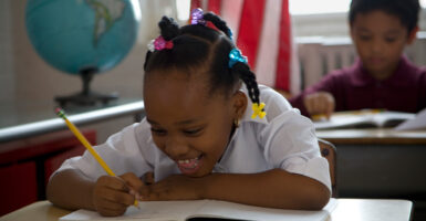 A girl write in a workbook at her desk at school with an American flag and world globe behind her.