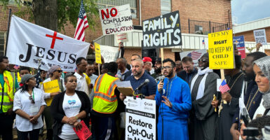 A group of protestors are gathers around a podium where a man in blue Muslim garb speaks. The group is outdoors and there are trees and a brick building in the backround.