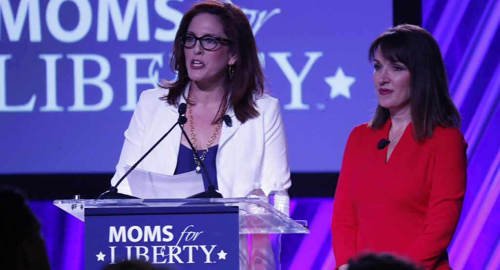 Tiffany Justice in a blue dress and white blazer speaks with Tina Descovich in a red dress in front of a Moms for Liberty logo