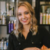 Young blonde hair stylist smiles in front of hair products