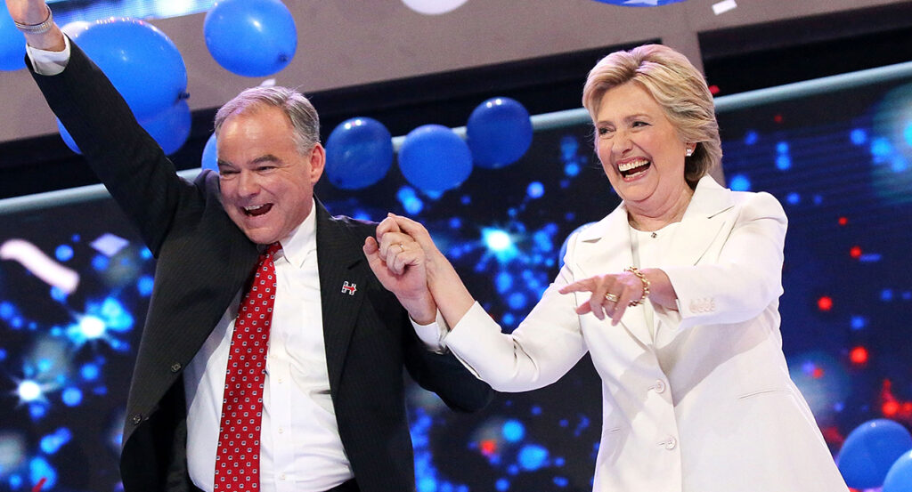 Tim Kaine in a black suit holds hands with Hillary Clinton in a white suit as they laugh and smile.