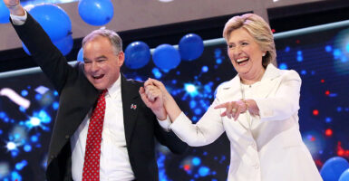 Tim Kaine in a black suit holds hands with Hillary Clinton in a white suit as they laugh and smile.