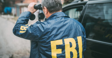 The back of a man's jacket with the letters "FBI" printed on it. From behind, he can be seen using binoculars and is standing next to a car.