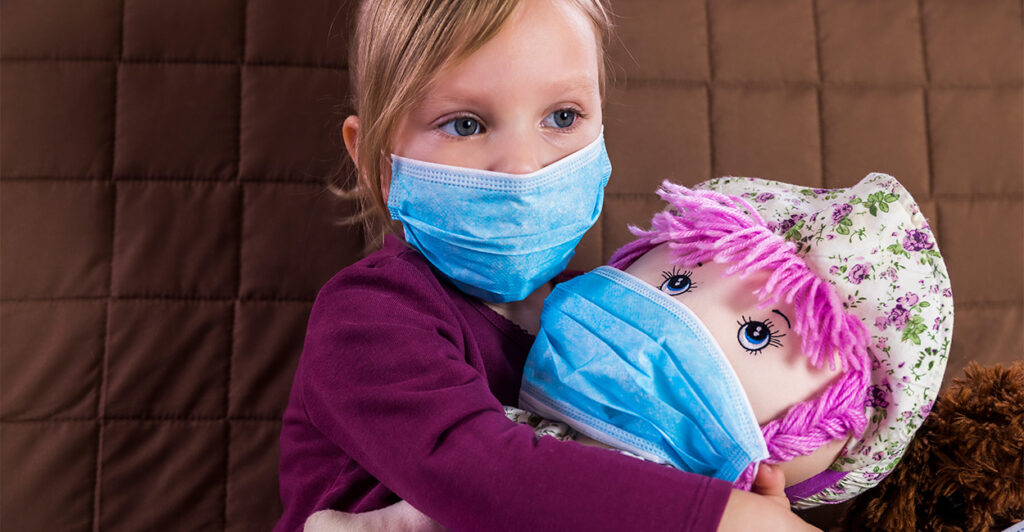 Girl in blue mask holds pink-haired doll.