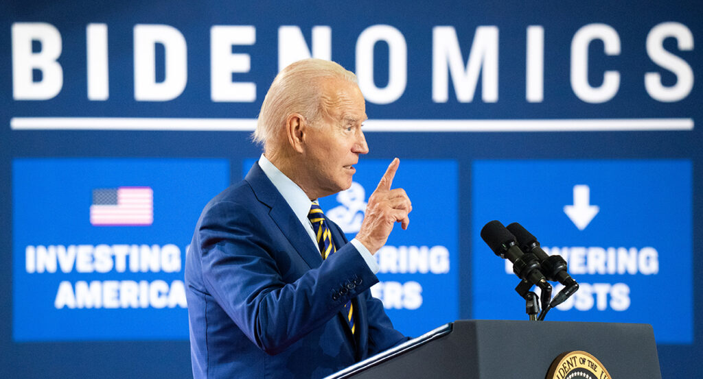 Joe Biden in a blue suit in front of a sign reading "Bidenomics"