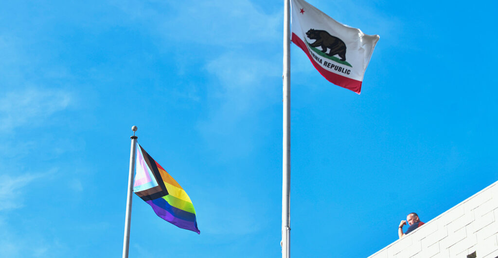 A pride flag and California flag are seen flying on a sunny day.