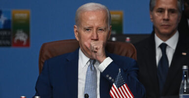 Joe Biden in a blue suit coughs with Antony Blinken looking over his shoulder