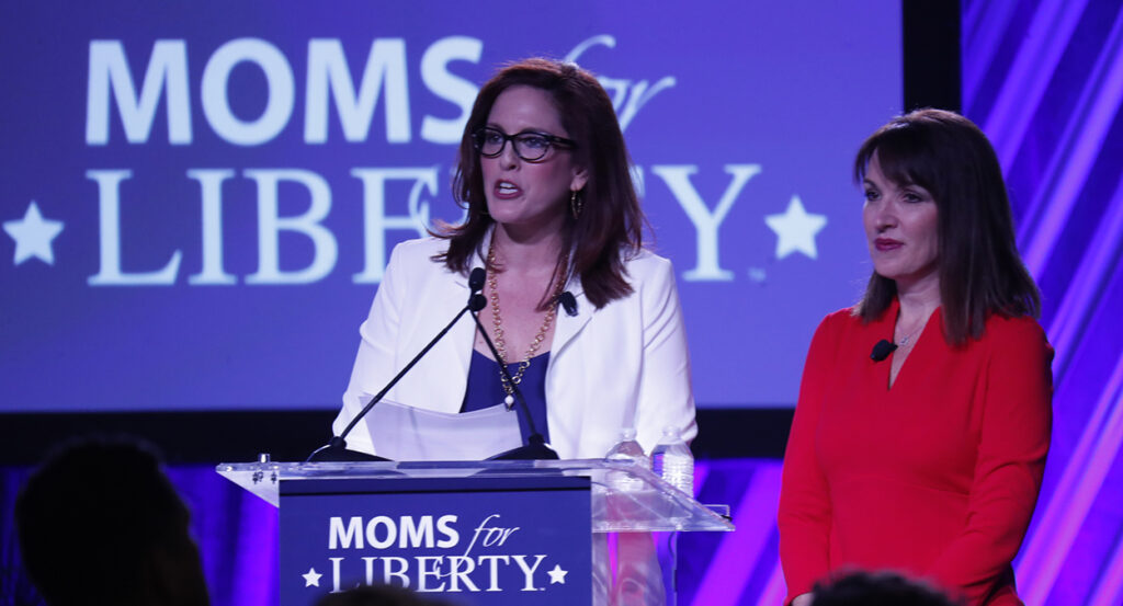 Tiffany Justice in white and Tina Descovich in Red in front of a Moms for Liberty banner