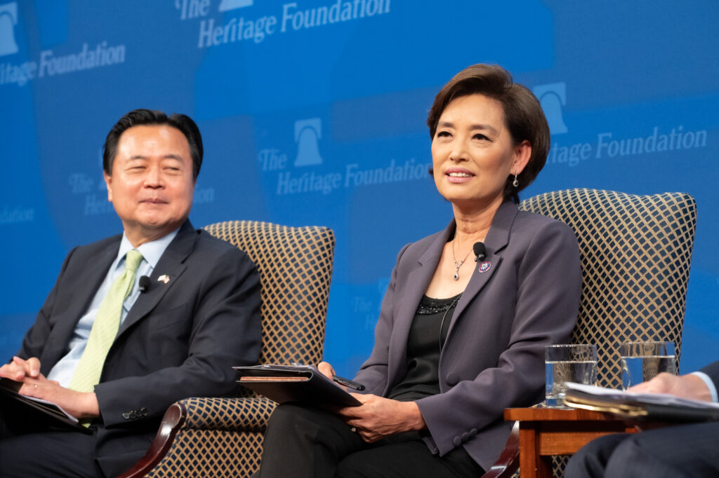 An asian man and woman sit side by side in business clothes and talk. They sit in front of a blue background.