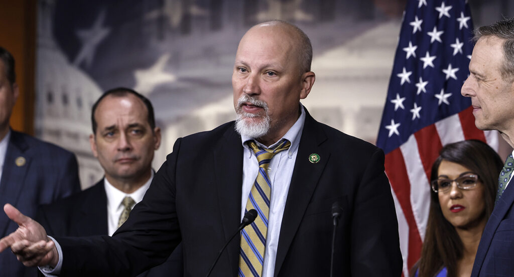 Chip Roy, bald and with a goatee, gestures in a black suit with a multi-colored striped tie