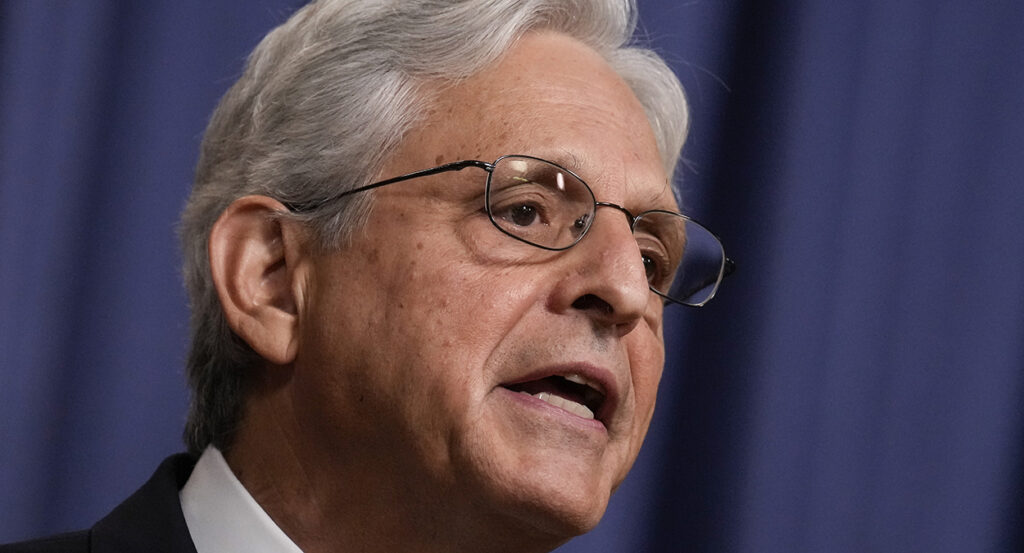 Merrick Garland with white hair in a suit and wearing glasses stands in front of a blue background