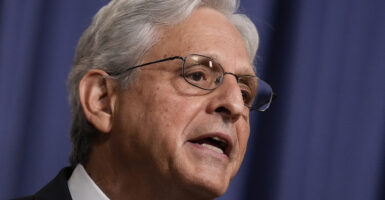 Merrick Garland with white hair in a suit and wearing glasses stands in front of a blue background