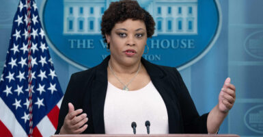 Shalonda Young in a white shirt and black jacket speaks in front of an American flag and the White House logo