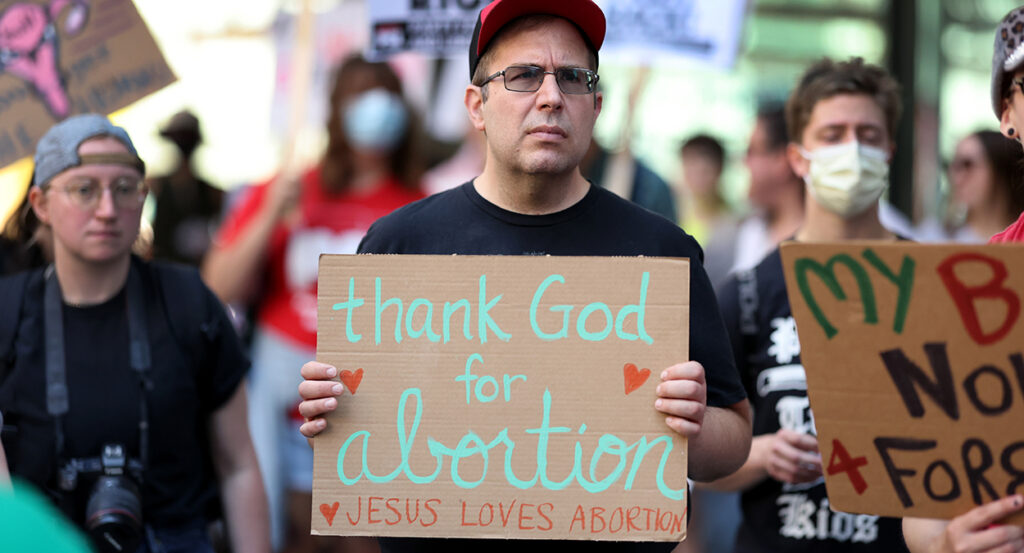A man in a black shirt holds a sign reading "Thank God for abortion"