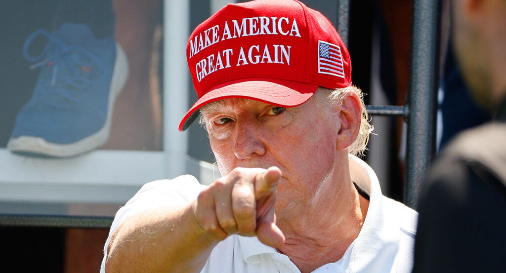 Donald Trump, wearing a "Make America Great Again" hat, points at the camera.