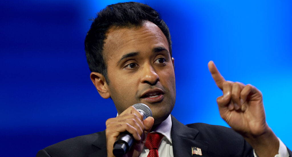 Vivek Ramaswamy raises a finger while speaking in the microphone and wearing a suit with an American flag pin.