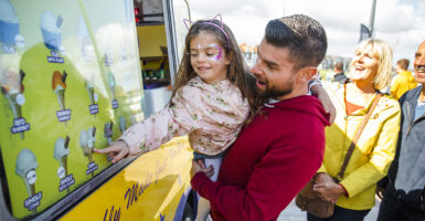 a man carrying his daughter while she is pointing at an ice cream menu on an ice cream truck