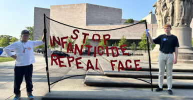Two men stand in front of a tan building outdoors holding a net with letters that spell the phrase "Stop infanticide, repeal FACE."