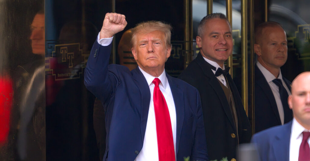 Donald Trump wears a blue suit jacket and a red tie. He holds up his fist.